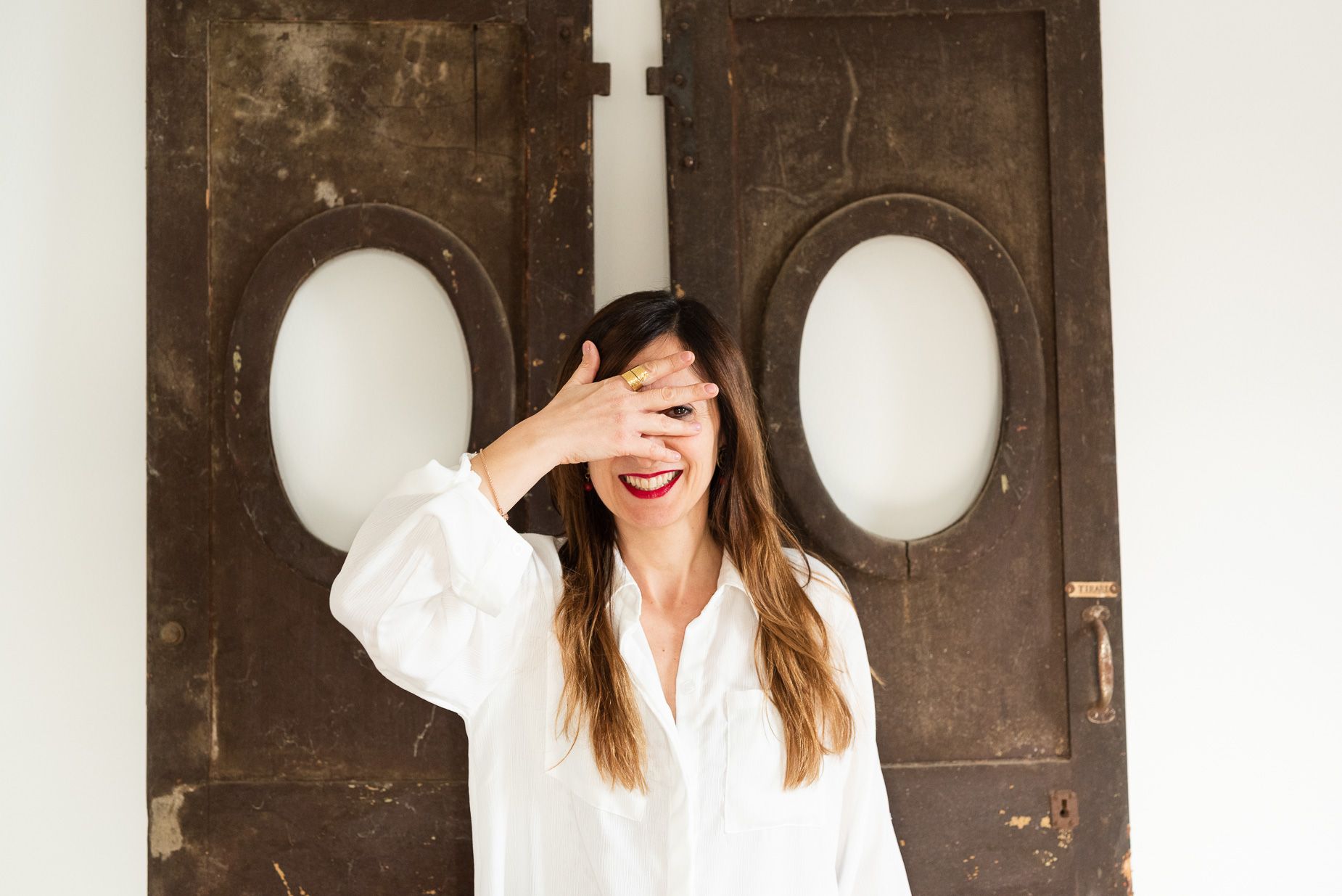 Donna sorridente con camicia bianca e rossetto rosso davanti a un portone di legno